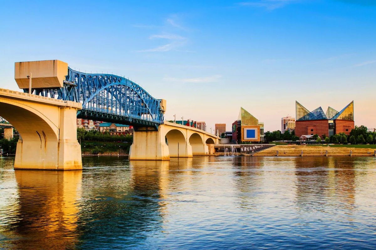 A serene riverside scene shows a blue bridge stretching over calm waters, modern city buildings in the distance, and the tranquil reflection of the river. The ambiance suggests opportunities for strolling, biking, and enjoying local outdoor culture.