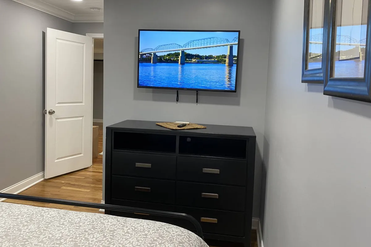 A spacious primary bedroom featuring a comfortable bed with plush bedding, a dresser topped with decorative accents, a smart TV displaying a scenic bridge over water, and a door leading to a connected space. The calm lighting creates a restful atmosphere ideal for winding down.