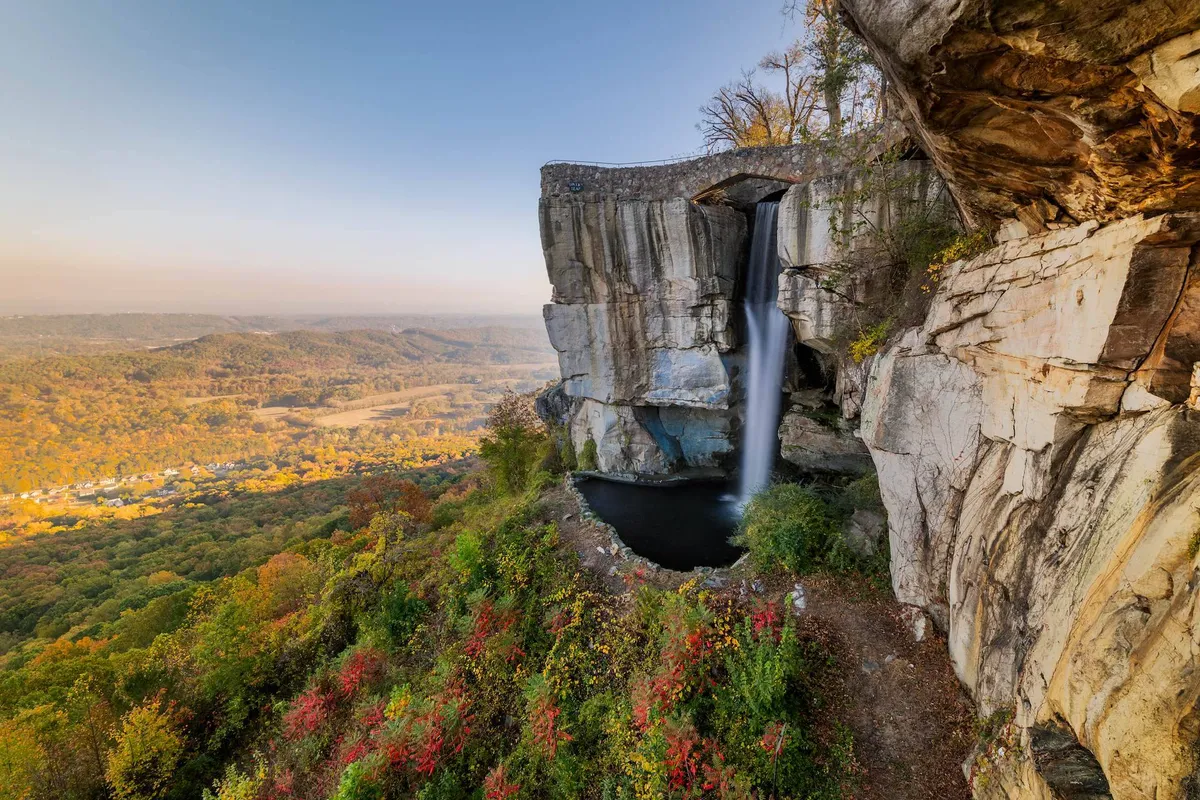 A dramatic natural landscape featuring a cascading waterfall, unique rock formations, a sweeping valley vista, and colorful autumn foliage. The scene evokes a sense of outdoor adventure and natural discovery.