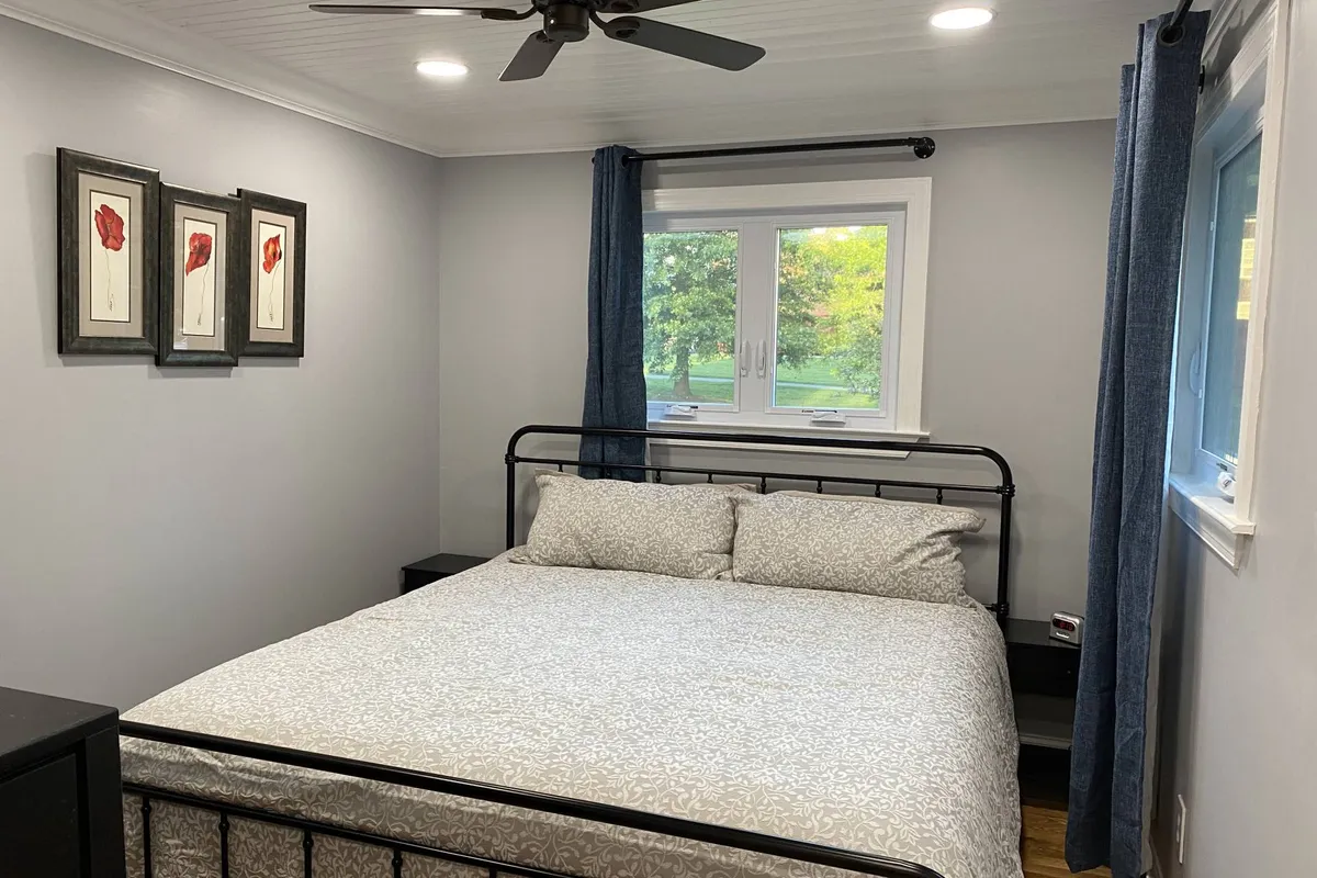 A peaceful bedroom scene featuring a cozy bed, soft bedding, a sunlit window framed by curtains, and subtle artwork. The ambiance is tranquil and inviting, ideal for unwinding after a busy day.