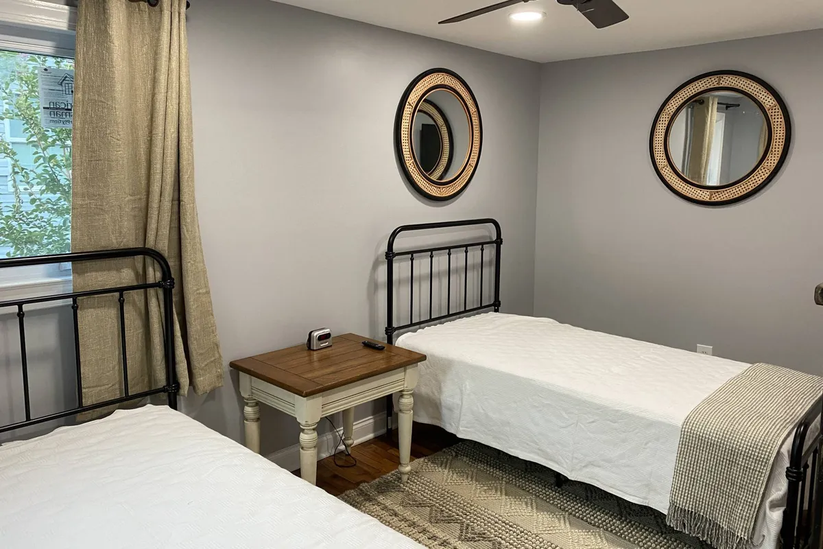 This image features two metal-framed beds set against a serene backdrop, with round mirrors above the headboards and a wooden bedside table between them. The room exudes a calm and cozy atmosphere, perfect for unwinding after a long day. The subdued lighting and soft textures create a welcoming environment conducive to both rest and quiet reflection.