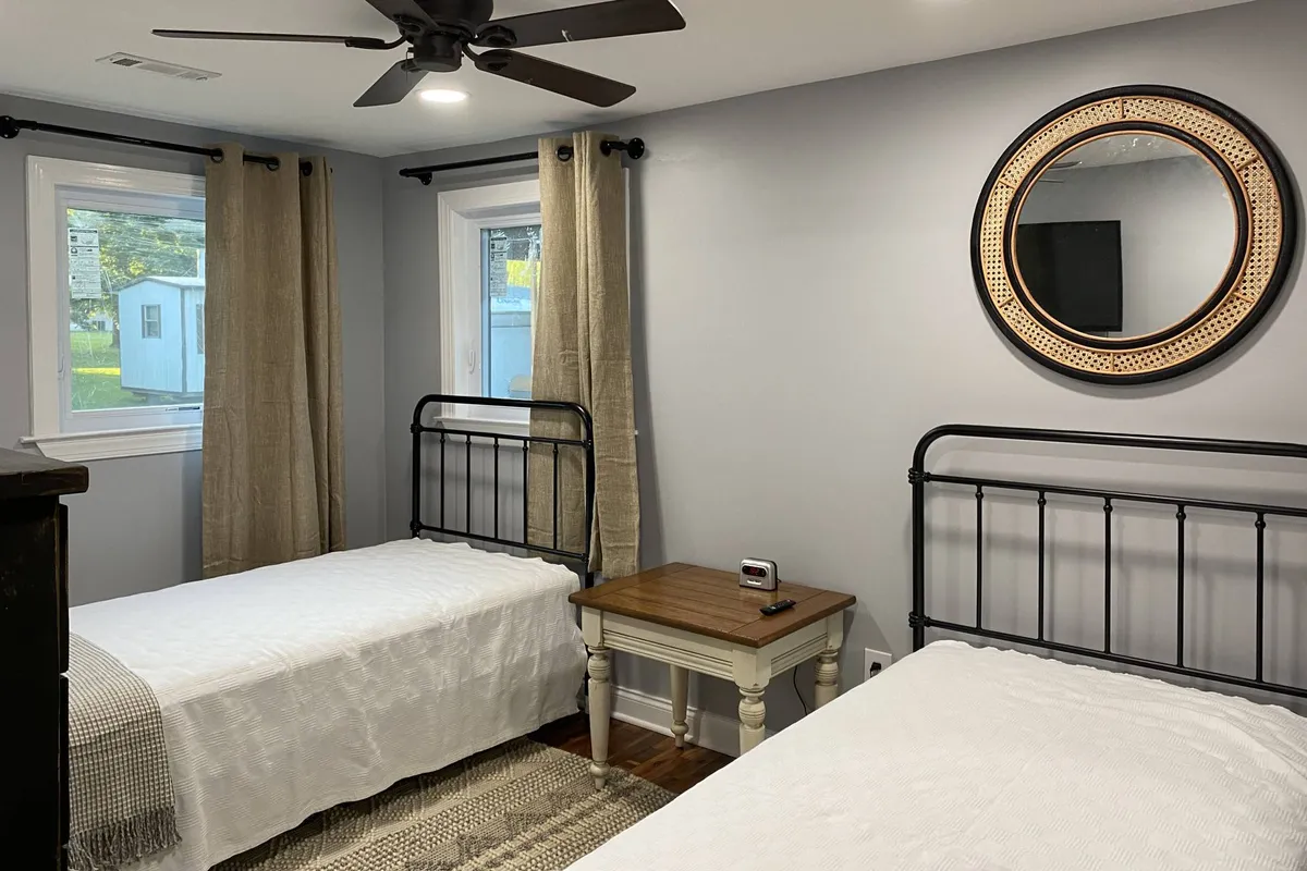 A cozy twin bedroom with two metal-framed beds, soft bedding, a wooden nightstand, and a decorative round mirror. The serene ambiance is perfect for children or guests needing flexible sleeping arrangements, and the room feels inviting for quiet mornings or restful nights.