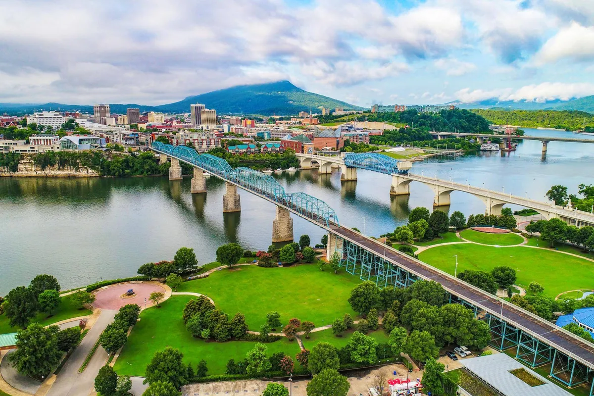 A vibrant view overlooking the river, iconic blue bridges, downtown Chattanooga’s skyline, framed by distant mountains. The scene is active with possibilities for walking, biking, picnicking, and family exploration along the waterfront.